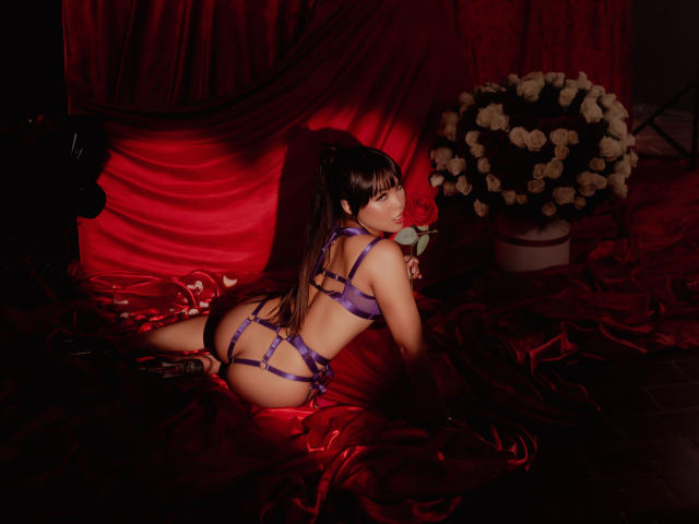 Heyly with long brown hair, wearing purple strappy lingerie, lying on red satin, holding a red rose, butt up, black heels, white rose bouquet on right.