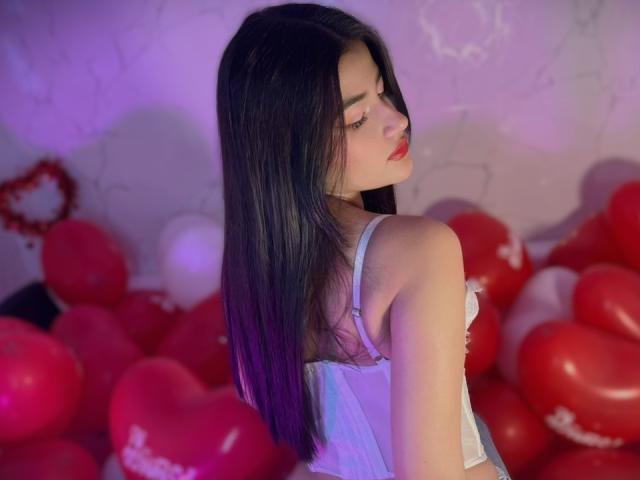 IrisBlair with long black hair, wearing a white lace bra, looking over her shoulder, surrounded by red and pink heart-shaped balloons, her red lips and eyelashes visible.