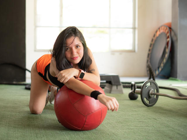 AbbyJamess, a woman with medium-length black hair, is on all fours on a green gym mat. She's wearing an orange and black sleeveless top and a black wristband. She's looking at the camera with a slight smile, resting her arms on a red exercise ball. A weight set and a trampoline are in the background. Natural light from a window illuminates the scene. She has a small tattoo on her left upper arm.