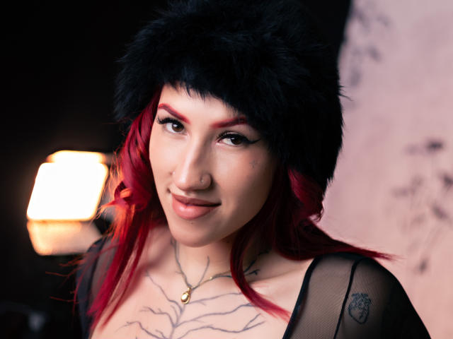 GingerxFoxy with red and black hair, black furry hat, nose ring, and tattoos, smiling with tongue out, wearing sheer black top, soft lighting, warm glow from lamp.
