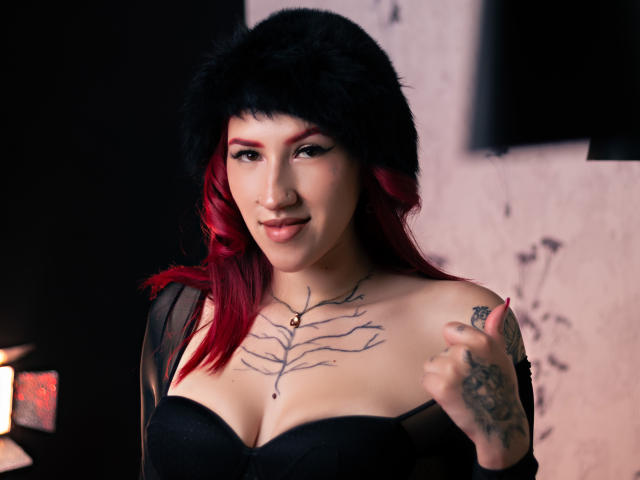 GingerxFoxy com cabelo ruivo, chapéu de pele preta, top preto sem alças, tatuagens no peito e no braço, sorrindo, segurando o top, mostrando os seios e um piercing no nariz..
