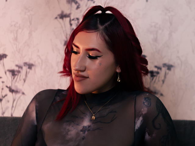 GingerxFoxy met rood haar, een zwarte doorschijnende top, tatoeages, een neusring, eyeliner, gouden oorbellen, een ketting, kijkt naar beneden, een zachte glimlach, zacht licht, sexy, aantrekkelijk, subtiele make-up, zelfverzekerd..