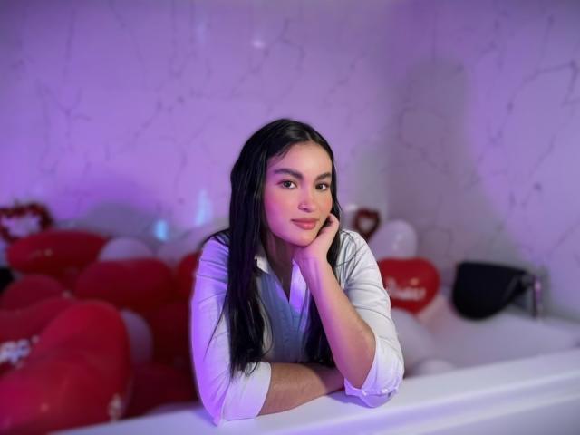 MiiaPalmer with long black hair, white shirt, leaning on white surface, head on hand, purple and pink light, red heart-shaped pillows in background, soft smile, relaxed.