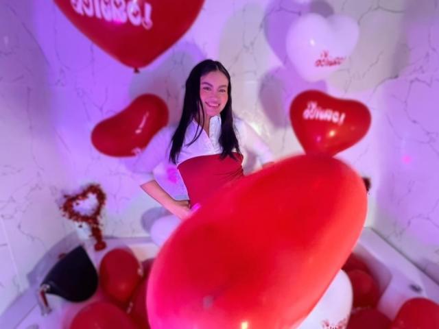 MiiaPalmer with long black hair, smiling, wearing a white shirt and red dress, surrounded by red heart balloons, sitting on a white chair in a purple-lit room.