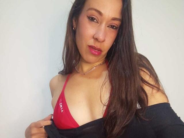 AmyRoberts with long brown hair, pink lipstick, and red bikini top showing small tits, black shirt off shoulder, silver necklace, and confident expression, against a plain white background.