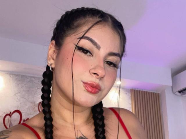 AlanaaWalker with puffy red lips, braided black hair, and a nose ring, wearing a red top, looking slightly to the side with a subtle smirk. Visible tattoos on her neck.