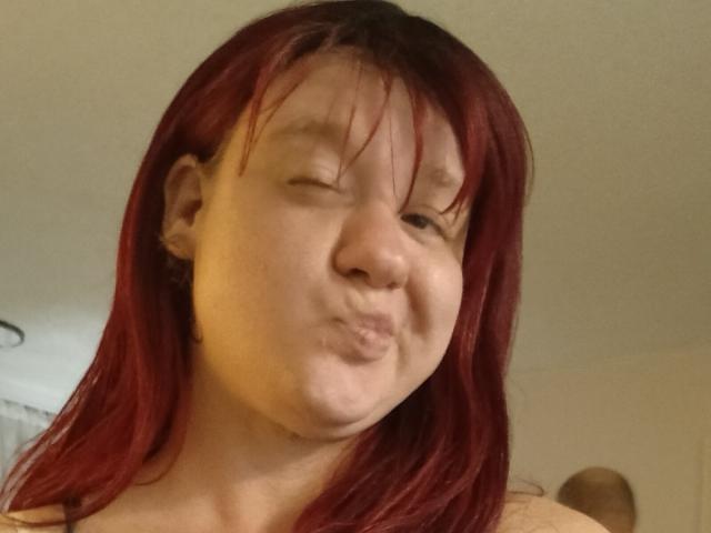 DeliciousRain with red hair, making a puffy lip face, slightly winking, showing a small nose, and looking cute, in a casual selfie. Her tits are not visible.