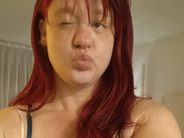 DeliciousRain with red hair, winking, and puckering her lips in a selfie. She's wearing a beige tank top, showing her freckled shoulders and small, perky tits.