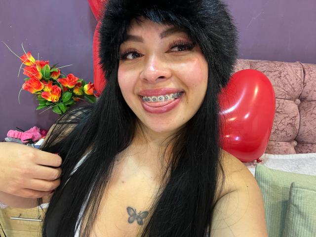 KateWelsin with black hair, wearing a furry hat, silver braces, tongue out, and a butterfly tattoo on her chest, smiling in a photo. Red balloon and flowers in background.