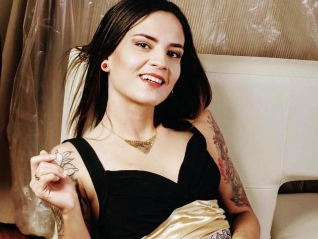 EmmaHerby with dark hair, black dress, gold necklace, smiling, holding a flower, tattoo on right arm, sitting on white chair, looking cute, showing her tits.