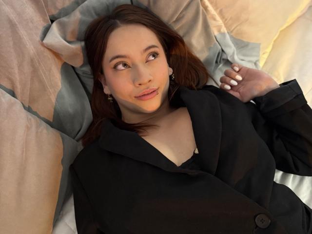 CleoMoon with brown hair, black blazer, and earrings, lying on a bed, looking to the side, showing a cute smile, with her hand near her face.