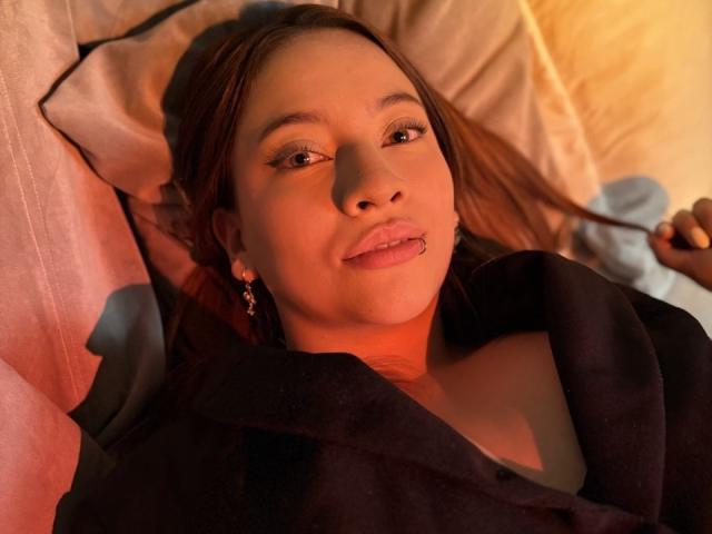 CleoMoon lying in bed, brown hair, slight smile, pink lips, nose piercing, black blanket, white pillow, warm light, subtle makeup, earrings, soft texture, relaxed expression.