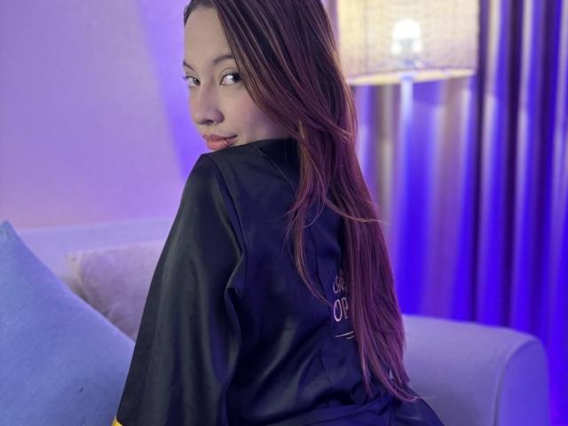 CleoMoon with long brown hair, wearing a black silky robe, looking over her shoulder, sitting on a white sofa, blue pillow, purple and blue lighting, subtle smile.