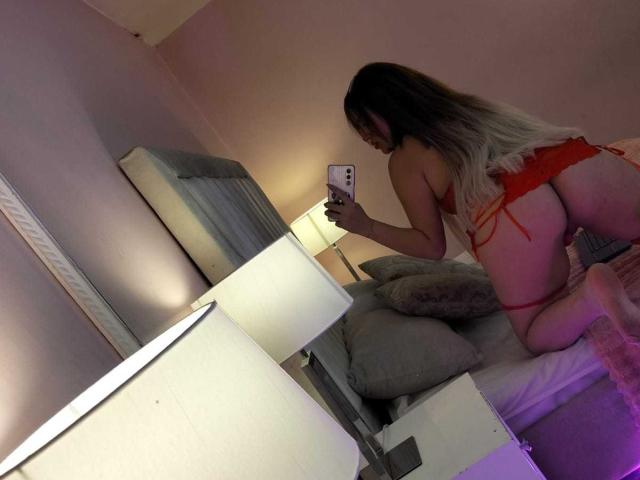 ZoeyClark in red lingerie, on knees, showing her ass, taking a selfie with a phone, long blonde to brown hair, in a dimly lit bedroom with a bed and lamp.