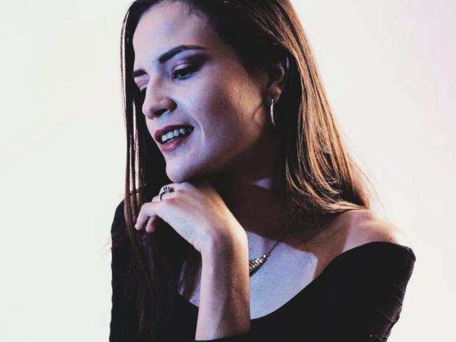 EmmaHerby with long brown hair, smiling, wearing an off-shoulder black top, holding her chin, subtle makeup, small hoop earrings, necklace, looking to the side, soft light.