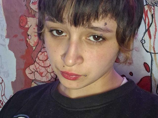 MichellJula has short brown hair, brown eyes, and a nose piercing. She also has a lower lip piercing and is wearing a black shirt. Her face is slightly freckled.