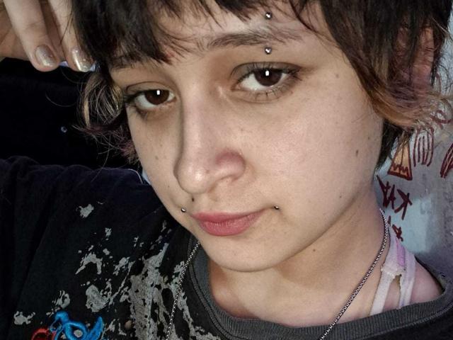 Close-up of MichellJula with short, dark brown hair and brown eyes. She has small, silver facial piercings in her eyebrows and lower lip. Her skin is light with a few small blemishes. She's wearing a black, graffiti-style shirt with gray and white patterns, and a silver chain necklace. Her right hand is raised, showing gray-painted nails. The expression is neutral with slightly pursed lips. The image is a photograph.
