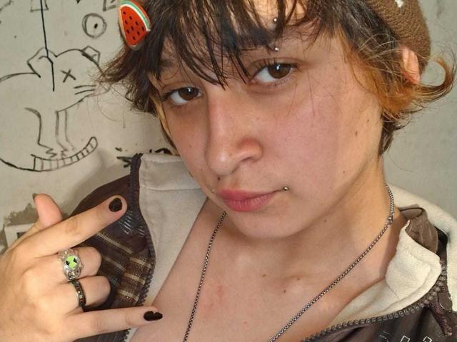 MichellJula has a pensive look with a nose ring, lip piercing, and a green ring with a red gem. Wearing a brown cap, brown jacket, and black nail polish.