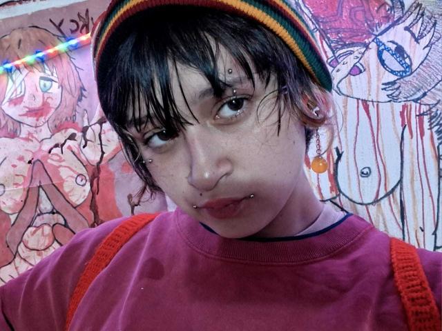 MichellJula has a short, messy black haircut with bangs and wears a rainbow-striped beanie. She has multiple facial piercings, including a nose ring and a lip stud, and small orange earrings. Her pink shirt has orange straps, and she has a blue choker. The background features colorful, abstract, sketch-like drawings of a person with blue eyes and a wolf-like figure. The image has a slightly blue-toned, digital photograph appearance.