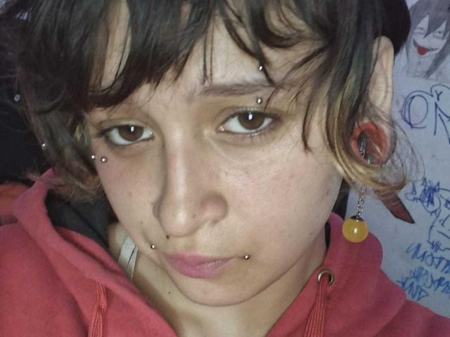 MichellJula has a pouty face, brown eyes, and multiple facial piercings: nose, upper lip, and above each eyebrow. Wearing a red hoodie with yellow earring on left.