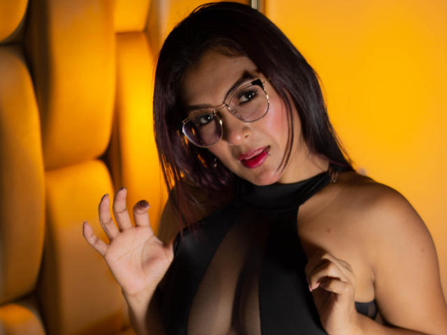 EvelynJohnsoon with dark hair, glasses, and red lipstick, wearing a sheer black top, exposing her tits, raising her hands, against a yellow, padded wall.