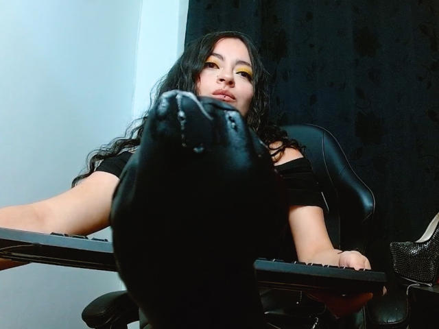 LunaTikka with long black hair, yellow eyeshadow, and black off-shoulder top, leaning on desk, black chair in foreground, slightly upskirt, looking at camera, serious expression.