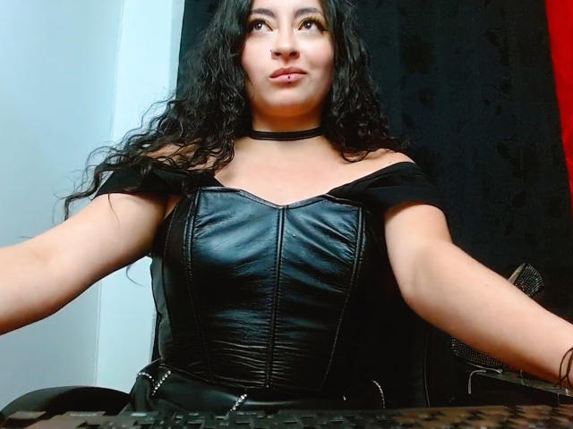 LunaTikka with curly black hair, wearing a black leather corset, choker, and off-shoulder sleeves, sitting at a desk, looking up with a slight smile. Boobs are visible.