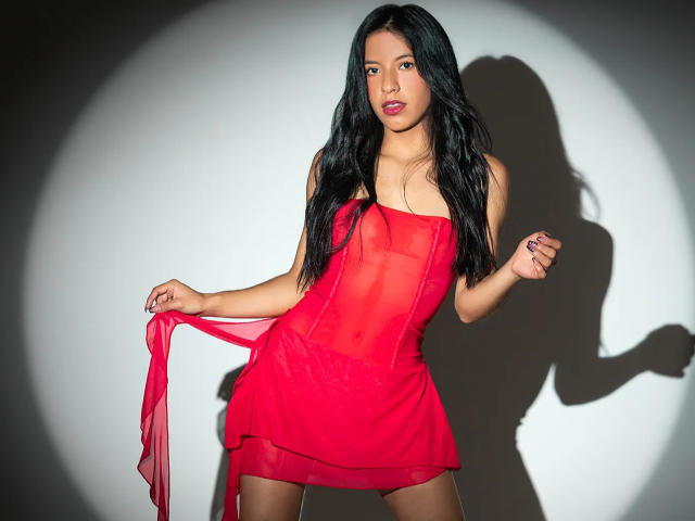 AlissaCavalli in a red sheer dress, holding a red ribbon, showing her ass and tits, with long black hair, standing, shadow on white wall, looking at camera.