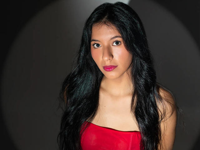 AlissaCavalli has long black hair, a red strapless top, and pink lipstick. She's looking straight at the camera with a neutral expression. Her tits are small and visible.
