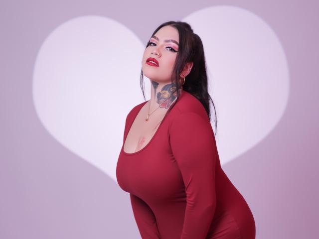 TessaMagic is a plus-sized woman with pale skin and dark hair in a ponytail. She wears a tight red long-sleeve top that accentuates her large breasts. Her makeup includes pink eyeshadow and red lipstick. A detailed tattoo of a skull and flowers covers her left neck and shoulder. She leans forward, looking at the camera with a slightly open mouth. A large white heart is projected behind her. The image has a soft, purple-gray background. Her expression is confident and slightly seductive. The photograph is realistic and well-lit.