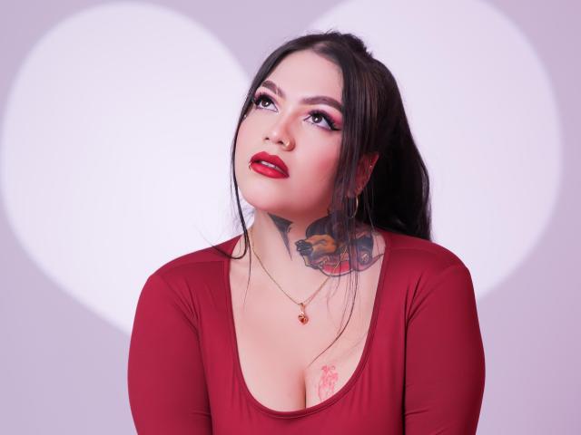 TessaMagic with red lipstick, dark makeup, and tattooed neck, wearing a red top, looking up with a slight smile, has a red pendant and a heart-shaped background.