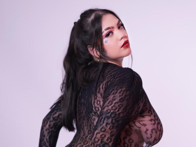 TessaMagic has dark lace top, showing her tits through the fabric, with red lipstick, black hair in a ponytail, and a small heart tattoo on her cheek.