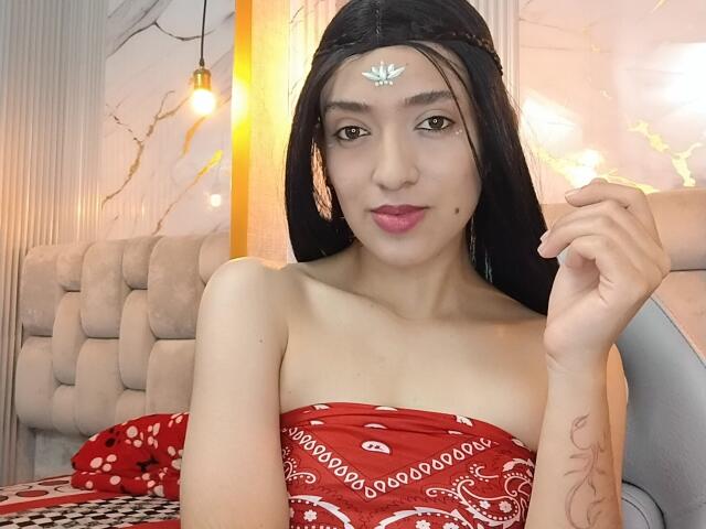 MichaelaMistress, an Asian woman with long black hair, wears a red strapless bandana top with white patterns. She has a small mole above her right lip and a tattoo on her left forearm. She touches her hair with her right hand, looking at the camera with a neutral expression. She has a silver forehead jewel. The background includes a beige tufted sofa and warm yellow light. The image is a photograph.