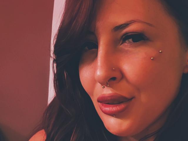 LinSyble has brown hair, a nose piercing, and two facial piercings above her lips. She's smiling with red lipstick and brown eyes, looking sexy.