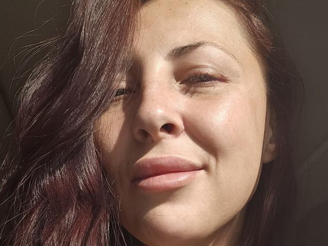 LinSyble's face with light skin, brown wavy hair, and subtle smile. She has pink lips, slightly squinted eyes, and a sunlit, natural look with visible skin texture.