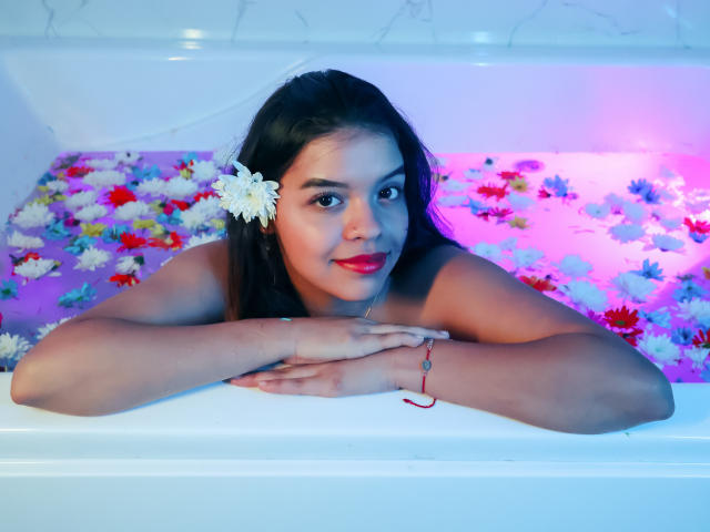 LiinaSantacruz, con cabello oscuro, una flor blanca en el cabello, lápiz labial rojo, acostada en una bañera llena de pétalos de flores de colores, con los brazos apoyados en el borde de la bañera, mirando a la cámara..