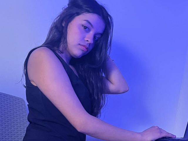 LiinaSantacruz, con cabello largo y oscuro, vestida con una camiseta negra sin mangas, está sentada con una mano en el cabello, mirando a la cámara, con iluminación azul, y el brazo derecho sobre una mesa..