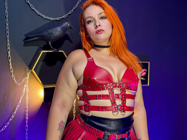 DominatrixMorrigan with bright orange hair, wearing a red leather harness, black choker, and red pleated skirt, showing her tits and a tattoo on her right arm. Black crow statue.