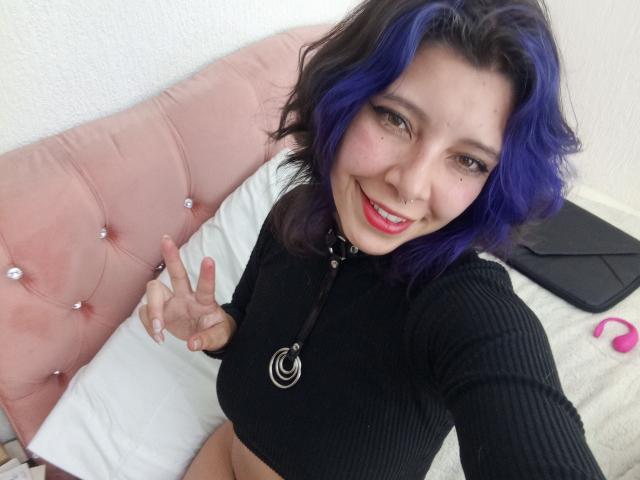 DarckVelvet with purple hair, smiling, black ribbed top, showing a peace sign, nose ring, and necklace with a crescent moon, sitting on a pink tufted bed.
