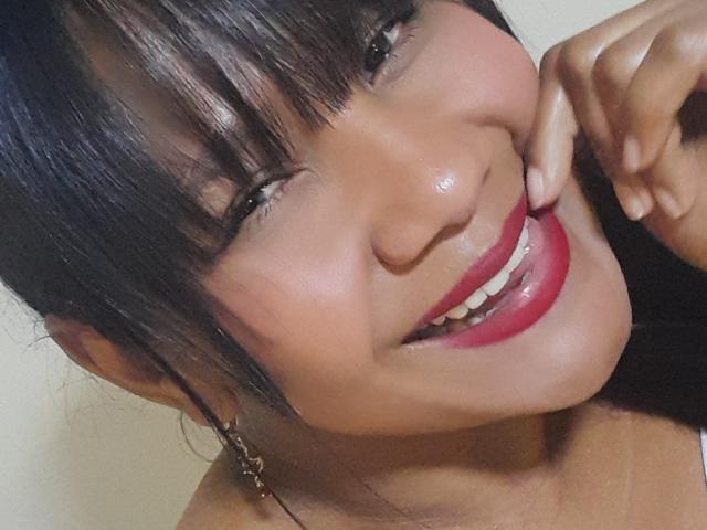 MarcelaHotty's face with dark straight bangs, glossy pink lipstick, and a shiny smile, touching her cheek with a hand, wearing small red earrings, has smooth tan skin.