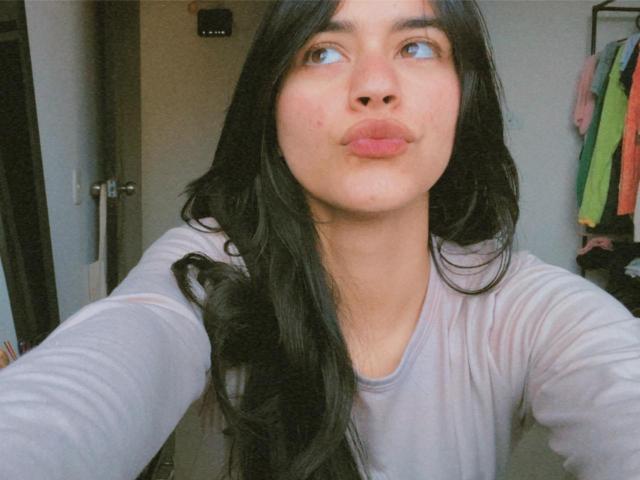 CharmingSofi with long black hair, pouty pink lips, and blue eyes, looking up, wearing a gray long-sleeve shirt, selfie-style.