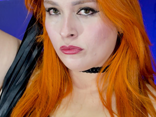 DominatrixMorrigan, con cabello naranja brillante, ojos verdes, delineador de ojos negro, lápiz labial rosa y un collar negro, frunciendo ligeramente los labios. Su piel pálida contrasta con su cabello rojizo..
