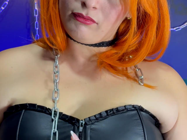 DominatrixMorrigan con i capelli arancioni vivaci, un top in pelle nera senza spalline e una collana a catena, che mostra il seno, indossa un collare nero e rossetto rosso..