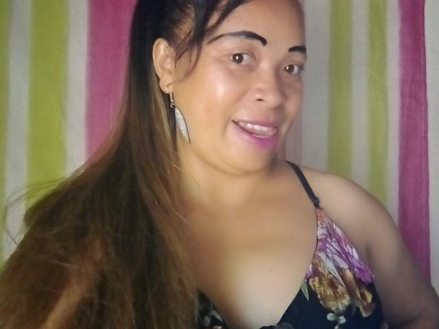 Close-up of Rahel6.9 with light brown skin and long brown hair in a ponytail. She has dark eyebrows, brown eyes, and pink lipstick. She wears a floral-patterned, black spaghetti strap top, and large white earrings. Her shoulders are visible, and she smiles slightly. The background has vertical stripes in pink, green, and white. The image is clear and focused.