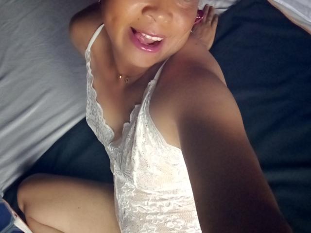 Rahel69 in white lace lingerie, smiling with pink lips, showing her tits and pussy, taking a selfie from above, dark bed background, visible arm and thigh.