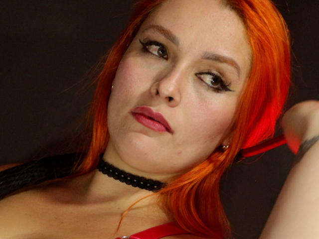 DominatrixMorrigan with bright orange hair, black choker, and red lipstick, staring with heavy black eyeliner, showing her tits in a red top. Dark background. Slightly puffy face.