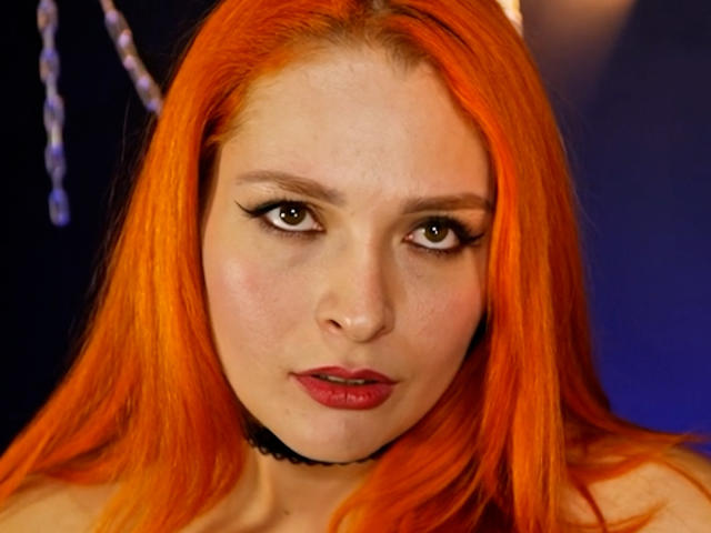 DominatrixMorrigan has bright orange hair, heavy eyeliner, and red lipstick. She's wearing a black choker and looking slightly annoyed with her eyebrows slightly raised.