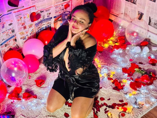 ArielleWilde, with black lace dress, kneeling on carpet, surrounded by red balloons, roses, and fairy lights, smiling with hands near face, showing her tits and ass.