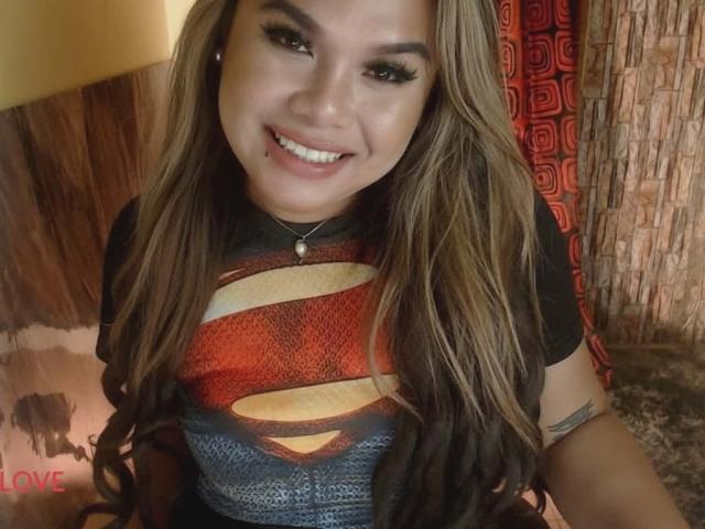 Kween has long, wavy brown hair with blonde highlights. She's smiling, showing white teeth. She's wearing a black Superman T-shirt with the iconic red and yellow "S" logo. She has a small tattoo on her right arm. Her makeup includes thick, dark eyeliner and long, curled eyelashes. She's wearing a silver necklace with a small pendant. The background includes a wooden wall and a red curtain with a geometric pattern. The word "LOVE" is visible in red text in the lower left corner.