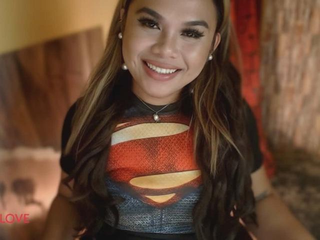 A portrait of Kween with long, wavy brown hair and light skin, smiling at the camera. Kween wears a Superman graphic t-shirt with a red, yellow, and blue Superman logo. She has dark, defined eyebrows, long eyelashes, and is wearing small pearl earrings. The focus is on her face and upper body. The background is blurred. The watermark "LOVE" is in the bottom left corner. The lighting is warm.